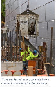 Three workers wearing bright yellow shirts move concrete into a column where it pours down a piece of metal.